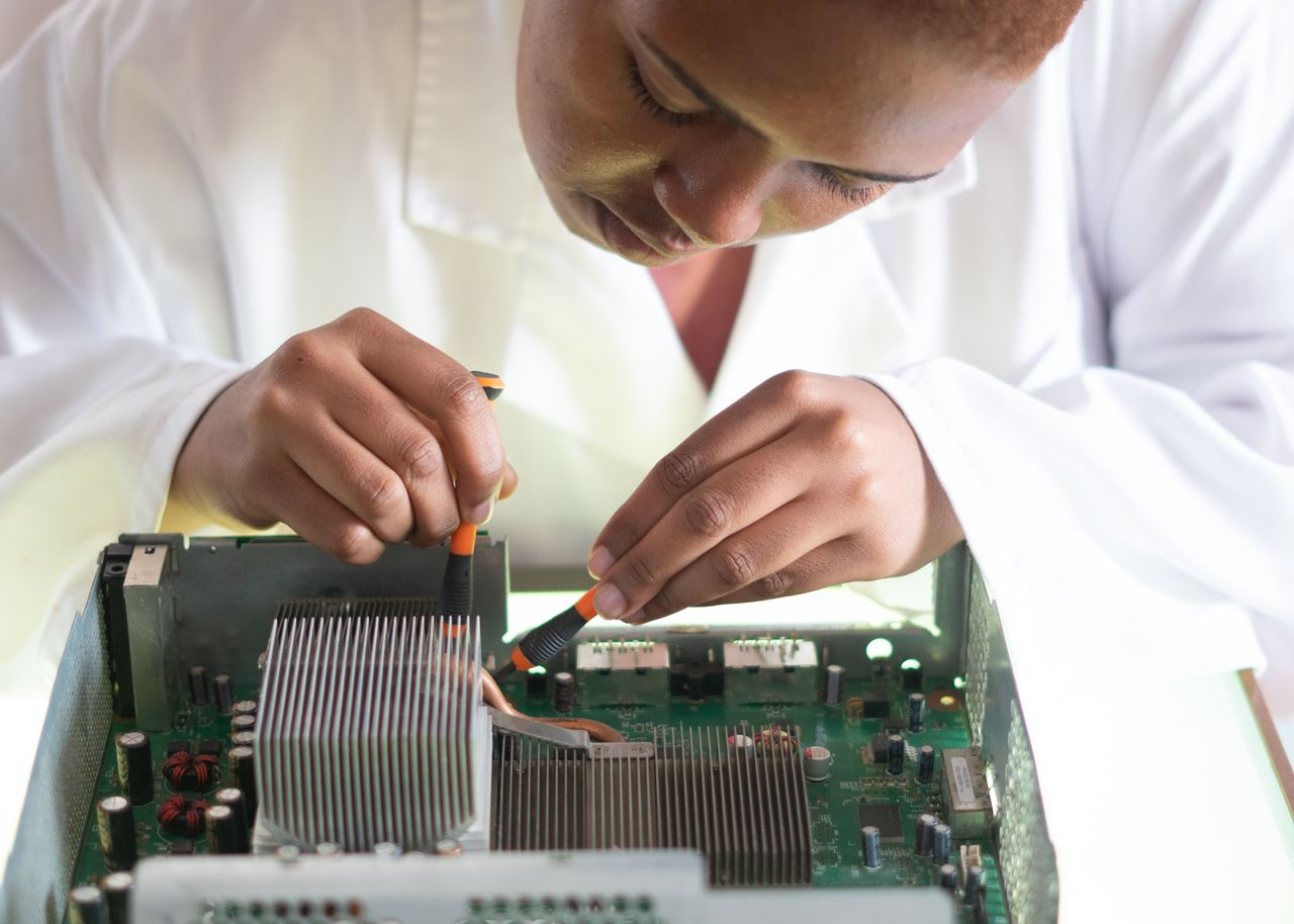 portfolio-01 Concentrated African American technician wearing lab coat and conducting expertise of motherboard by using screwdrivers while working in service center
